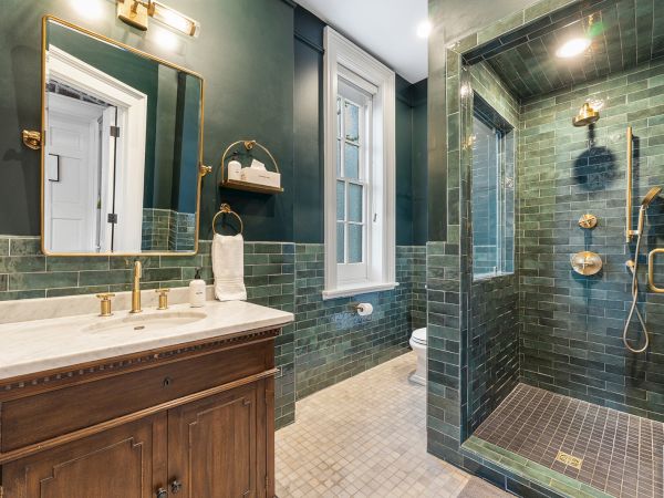 A modern bathroom with green tiled walls, gold fixtures, a vanity sink with mirror, and a walk-in shower stall.