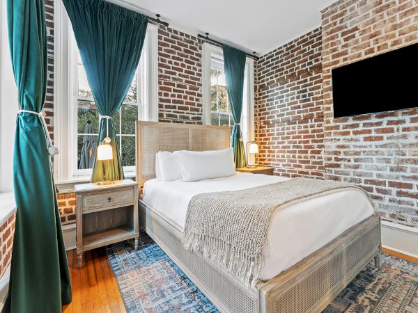 A cozy bedroom with a large bed, white linens, teal curtains, exposed brick walls, and warm bedside lamps.