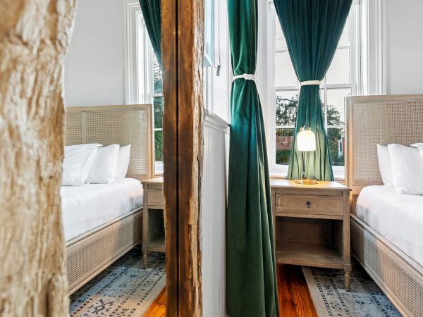 A cozy bedroom with two twin beds, white pillows, green curtains, a nightstand, and soft daylight through windows.