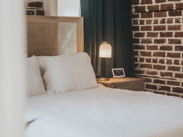 A cozy bedroom with a white bed, a nightstand with a lamp and photo frame, and walls with a brick pattern design.