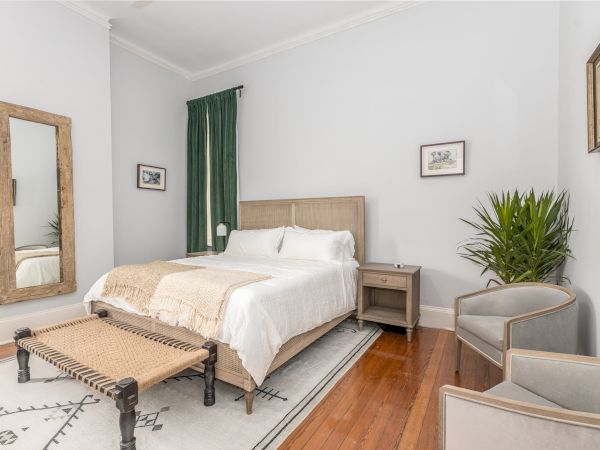A tidy bedroom with a bed, side table, large mirror, plants, green curtains, two armchairs, and framed artwork on the light grey walls.