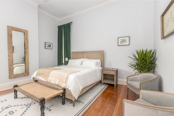 A tidy bedroom with a bed, side table, large mirror, plants, green curtains, two armchairs, and framed artwork on the light grey walls.