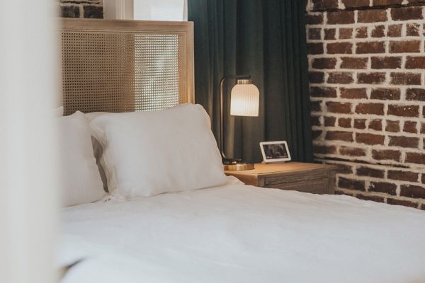 A cozy bedroom with a white bed, a nightstand with a lamp and photo frame, and walls with a brick pattern design.