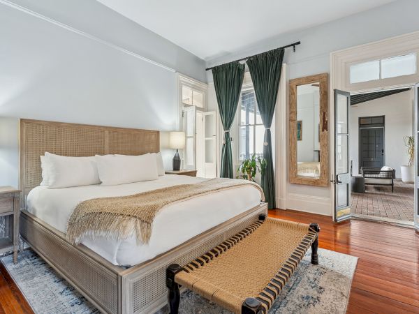 A stylish hotel bedroom with a large bed, beige textiles, green curtains, wooden floors, a bench at the foot, and a doorway leading to another seating area.