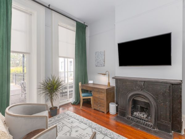 A cozy room features green curtains, a desk with a lamp, a wall-mounted TV, an ornate fireplace, a plant, and a pair of cozy chairs.