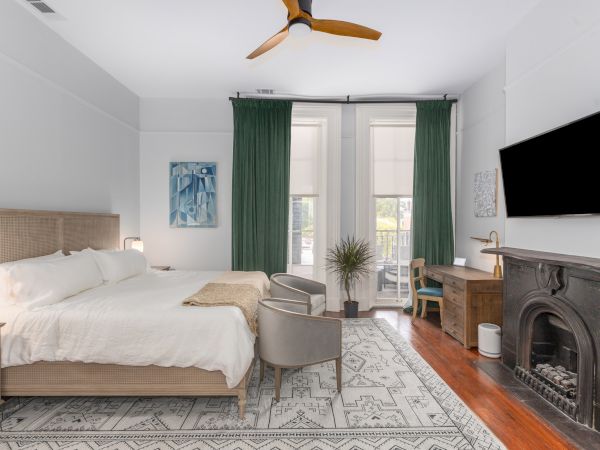 A cozy bedroom with a large bed, two chairs, green curtains, a fireplace, a wall-mounted TV, and a wooden desk with a plant.
