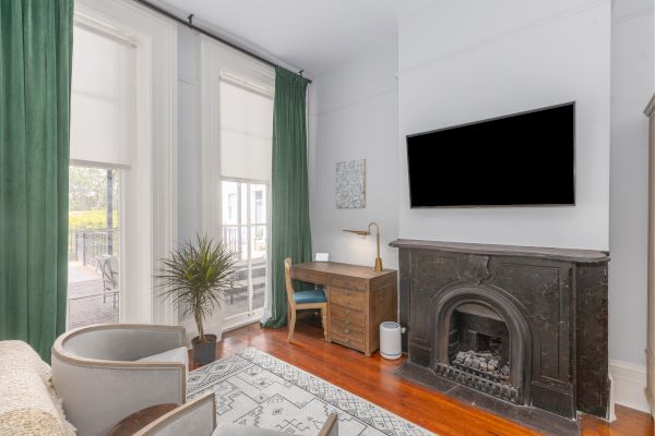 A cozy room features green curtains, a desk with a lamp, a wall-mounted TV, an ornate fireplace, a plant, and a pair of cozy chairs.