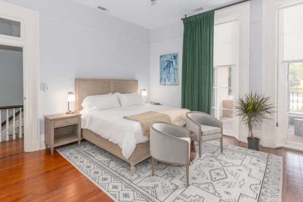 A cozy bedroom with a large bed, two bedside tables, two chairs, a rug, a green curtain, wall art, and a potted plant by a window.