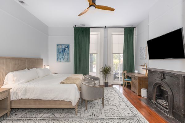 A cozy bedroom with a large bed, two chairs, green curtains, a fireplace, a wall-mounted TV, and a wooden desk with a plant.