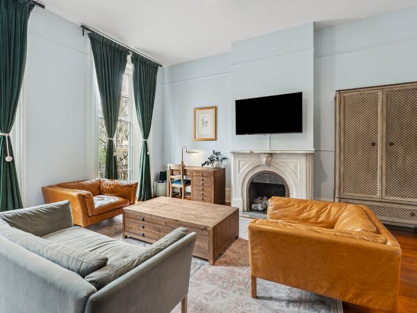 A bright living room with pale blue walls, two sofas (gray and brown), a wooden coffee table, a fireplace, wall-mounted TV, tall green curtains, and a wicker cabinet.