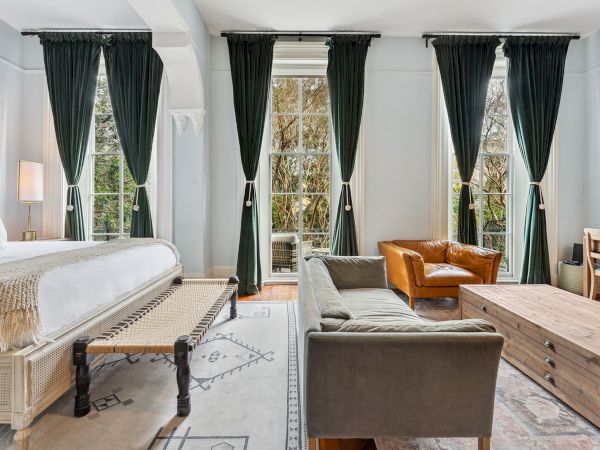 A bright hotel suite with a king bed, dark green curtains, a sitting area with gray sofa and brown chairs, a wooden coffee table, and a rug.
