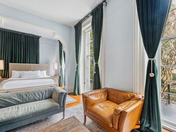 A stylish hotel room with a king bed, dark green curtains, a tan leather chair, a gray sofa, and large windows letting in natural light.