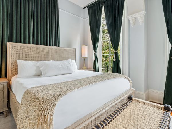 A luxurious bedroom with a large bed, white linens, beige headboard, soft lighting, and dark green curtains framing a bright window.