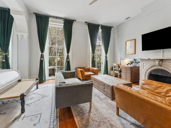A stylish living room with large windows, green curtains, a stone fireplace, modern sofas, a coffee table, and a mounted TV.