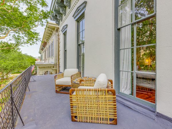 A sunny balcony with wicker chairs and white cushions, overlooking a leafy street, attached to a light-colored building.