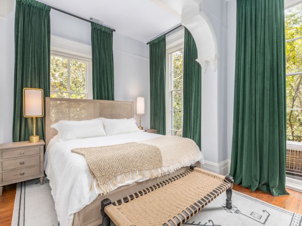 A cozy bedroom with green curtains, large windows, a double bed with neutral linens, bedside tables, and floor lamps on either side of the bed.