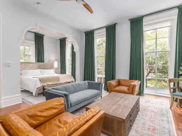 A spacious bedroom has a bed, green curtains, a couch, chairs, a coffee table, and a desk with a plant.