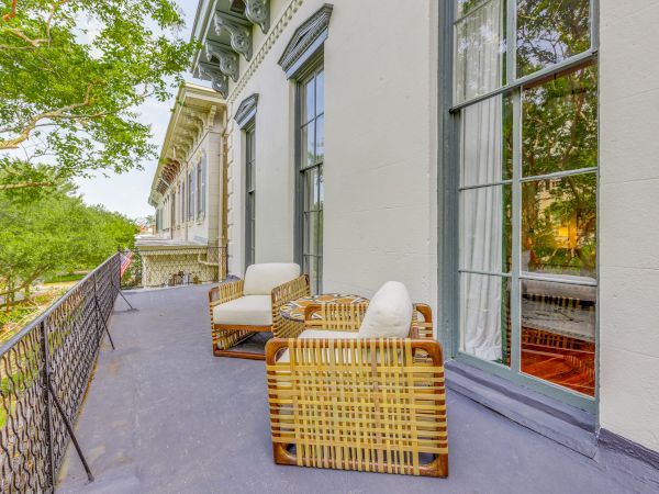 A balcony with two wicker chairs and cushions, iron railings, and a view of trees and neighboring buildings. The setting is serene and inviting.