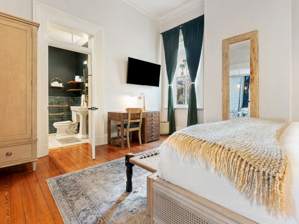 A cozy hotel-style bedroom with a white bed, beige throw, wooden dresser, desk and chair, wall mirror, rug, and an open doorway to a bathroom.