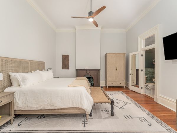 A well-lit bedroom with a large bed, wooden furniture, a ceiling fan, a TV on the wall, and an adjacent bathroom with an open door.