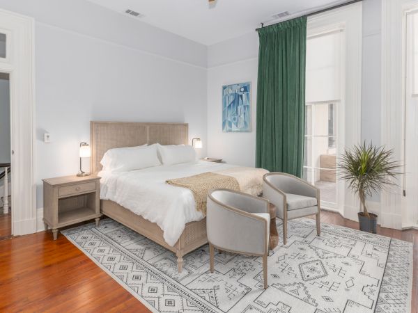 A bedroom with a large bed, two chairs, a small table, a potted plant, and a green curtain. The room features white walls and a patterned rug.