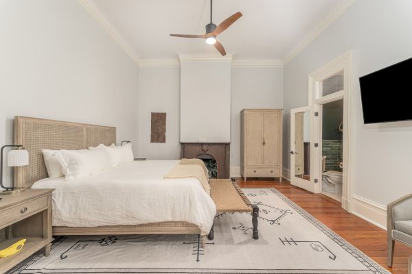 A well-lit bedroom with a large bed, wooden furniture, a ceiling fan, a TV on the wall, and an adjacent bathroom with an open door.