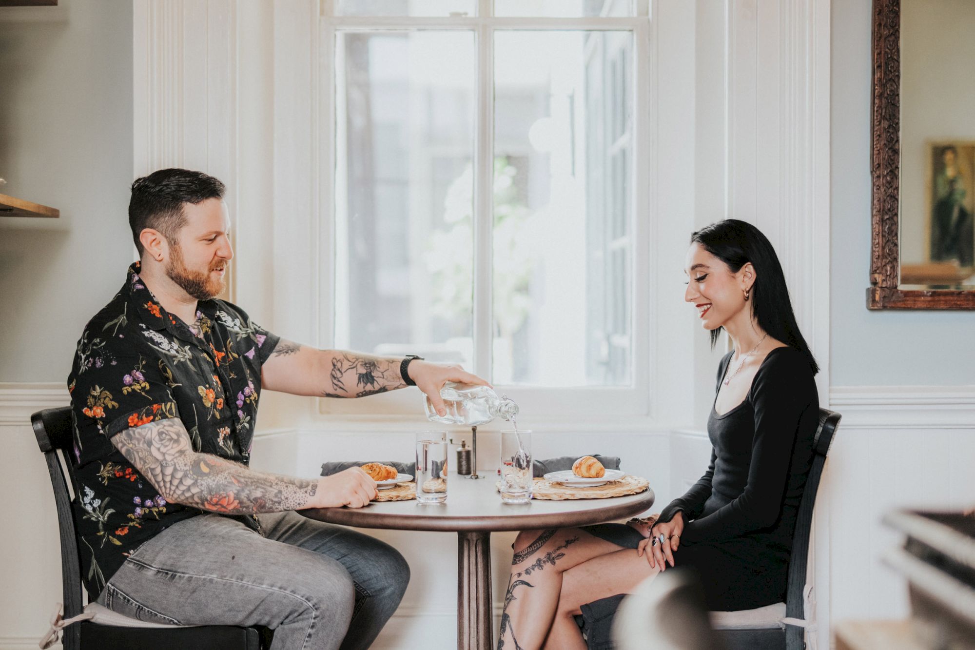 A man and woman sitting at a table in a bright room with a large window, engaged in conversation, and enjoying a meal together, with the man pouring water.