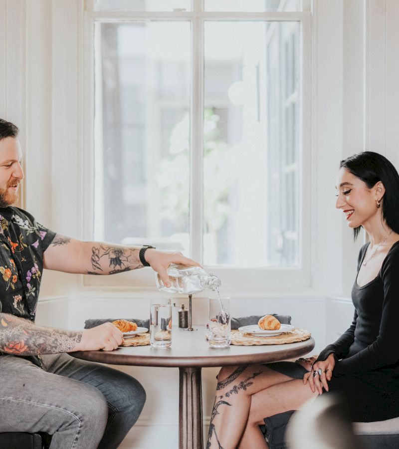 A man and woman sitting at a table with croissants and glasses, the man pouring water.