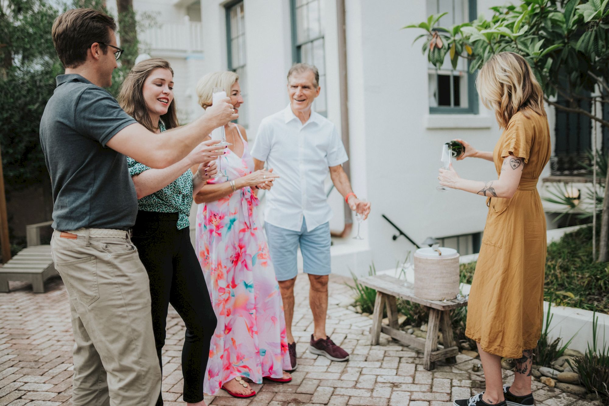 A group of people are gathered outdoors near a house, sharing drinks and enjoying each other's company, with one person pouring a drink from a container.