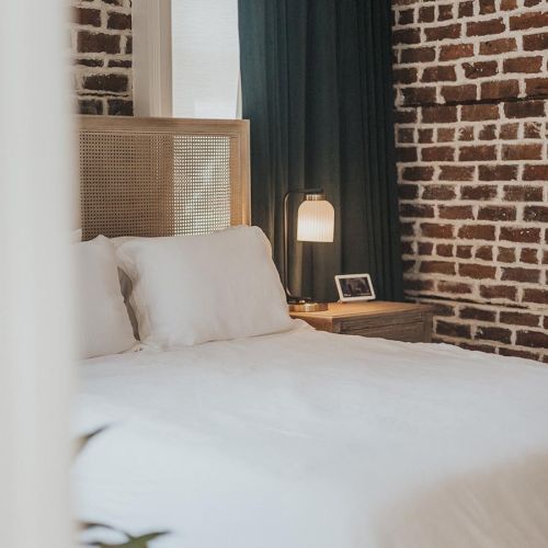 The image shows a cozy bedroom with a brick wall, white bedding, a bedside table with a lamp, and a small picture frame on it.