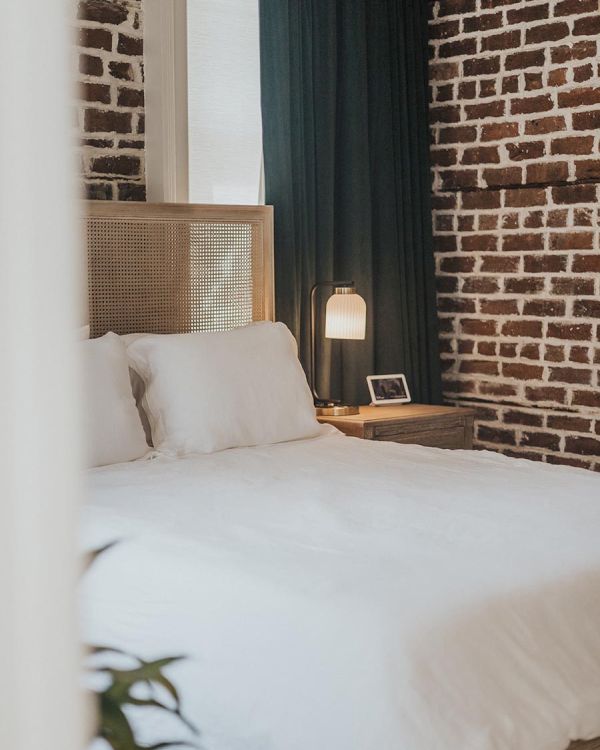 The image shows a cozy bedroom with a brick wall, white bedding, a bedside table with a lamp, and a small picture frame on it.