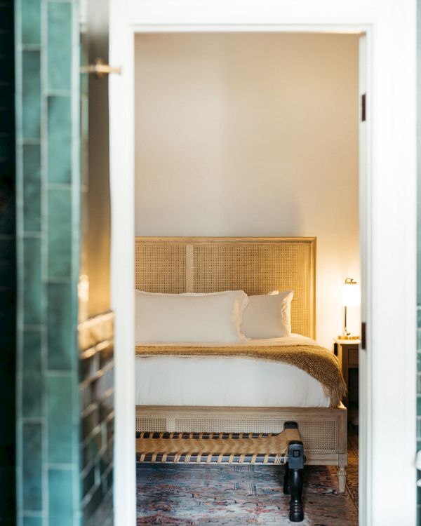 A cozy bedroom with a wooden bed, white bedding, a lamp on a nightstand, and a rug on the floor, viewed through a doorway.