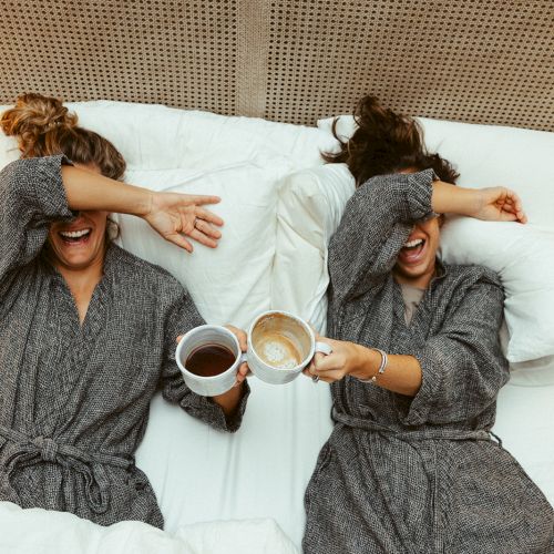 Two people in robes are lying on a bed, covering their eyes with their arms and holding mugs, appearing to enjoy a moment together.