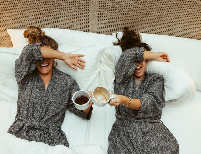 Two people in robes are lying on a bed, covering their eyes with their arms and holding mugs, appearing to enjoy a moment together.