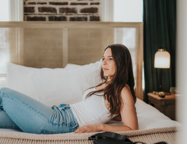 A woman in jeans and a white top lying on a bed with soft pillows, next to a lamp on a bedside table.