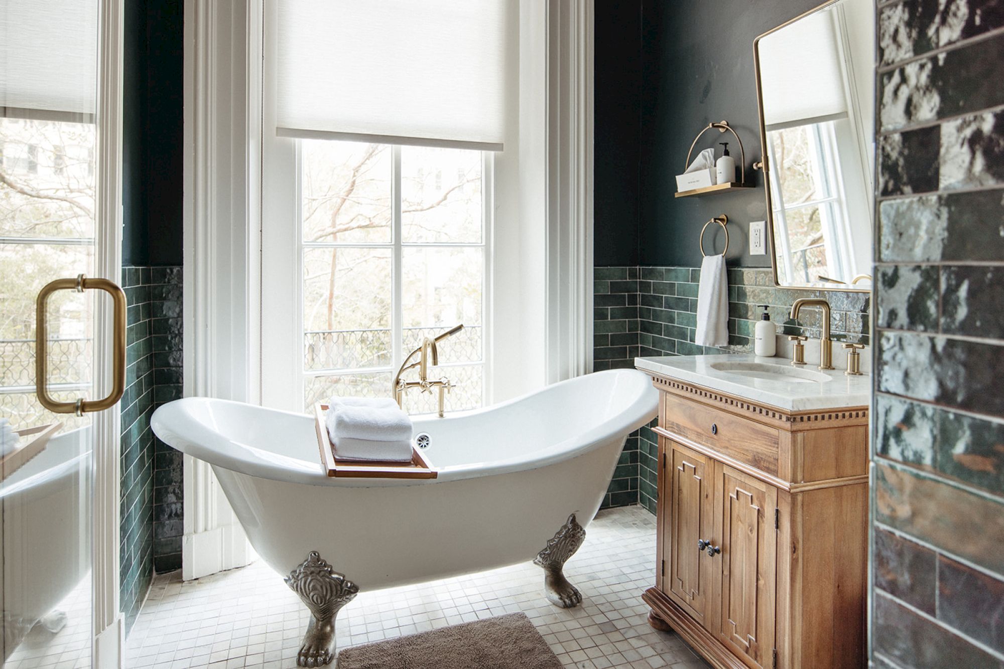 The image shows a bathroom with a clawfoot bathtub, wooden vanity, large mirror, and green tiled walls. Light streams through a window.