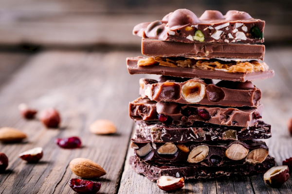 A stack of various chocolate bars with nuts and dried fruits, displayed on a wooden surface with some scattered nuts and cranberries around.