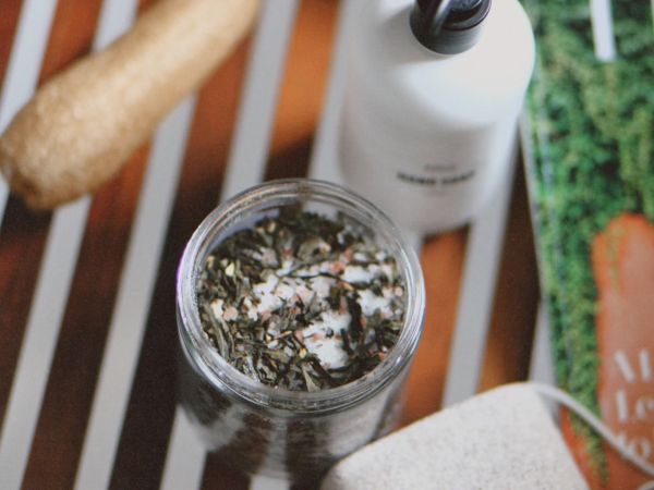 A wooden tray with bath items: a jar of bath salts, a lotion bottle, a pumice stone, a brush, and a partially visible green package.