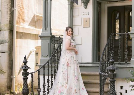 A smiling bride in a floral wedding dress stands on steps adorned with yellow and white flowers, outside an ornate building labeled 