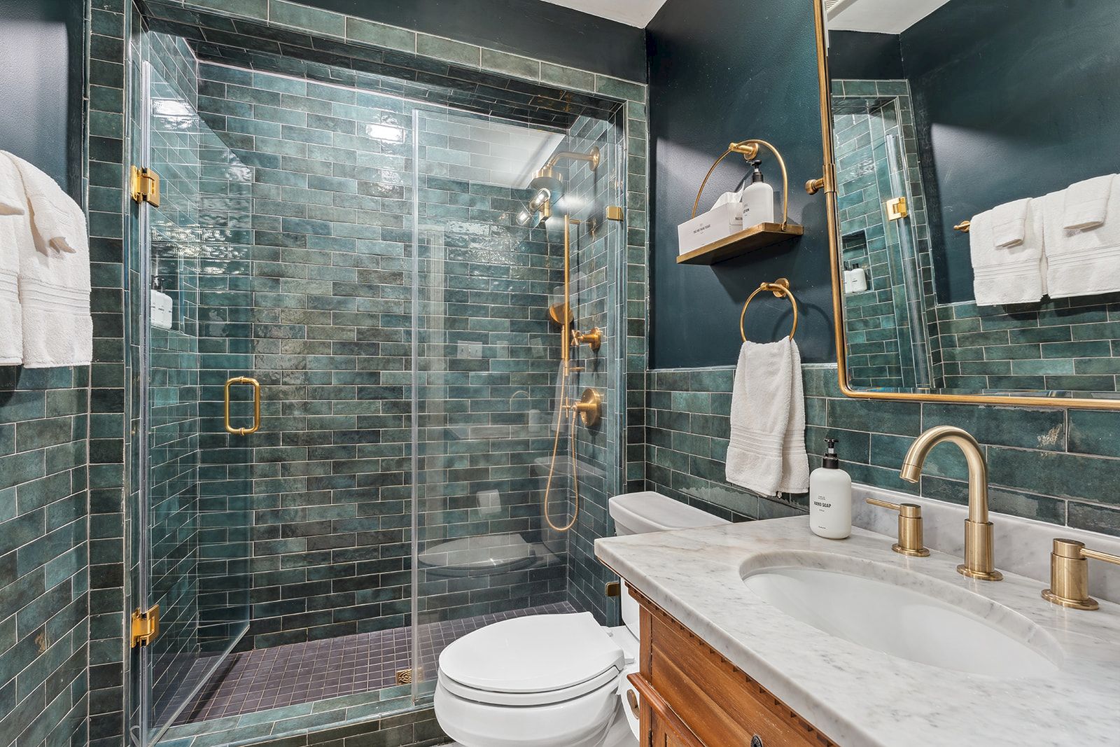 Modern bathroom with a glass shower, teal tiled walls, gold fixtures, towels, and a vanity sink with soap.
