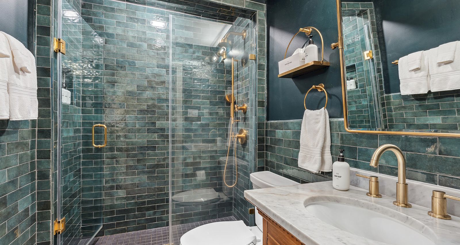 Modern bathroom with a glass shower, teal tiled walls, gold fixtures, towels, and a vanity sink with soap.