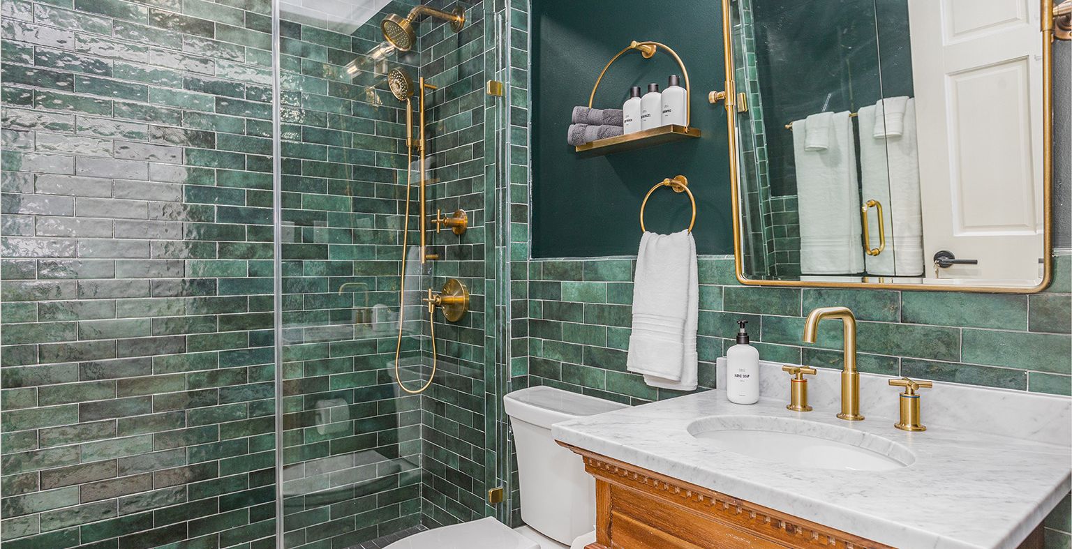A compact bathroom with a glass shower, green tile accents, wooden vanity, white countertop, and brass fixtures, plus a towel rack and shelves.