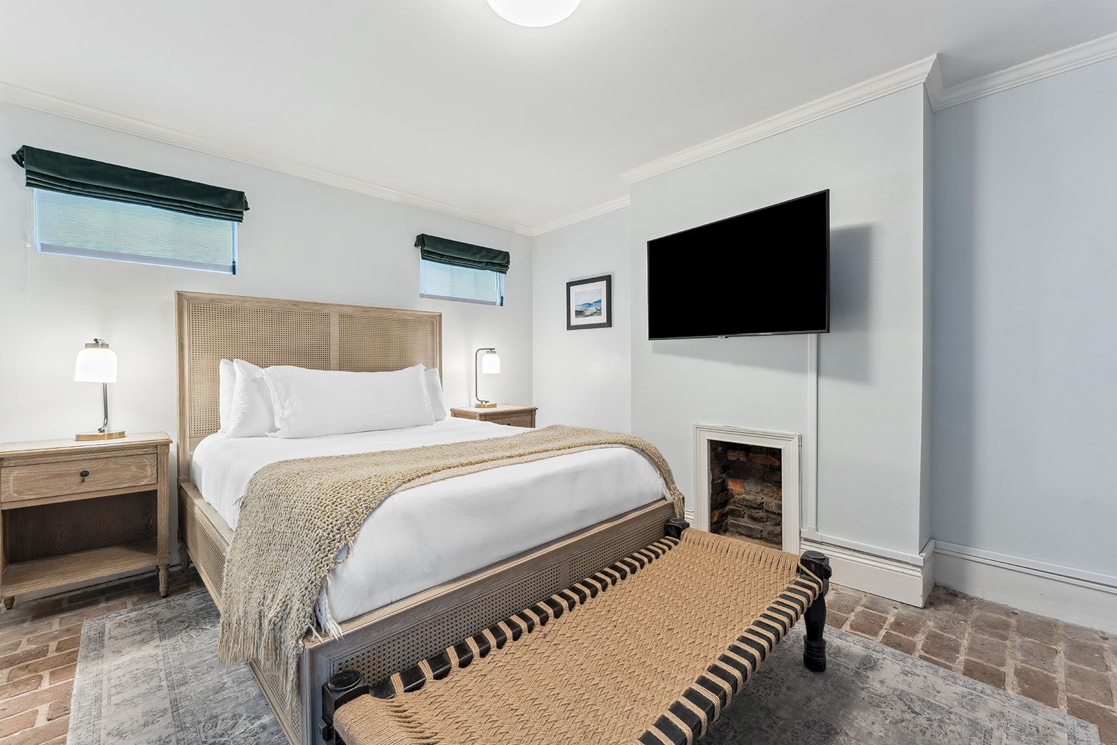 A bright hotel bedroom with a king bed, two nightstands, a bench at the foot, a wall-mounted TV, and a small fireplace-style area. End.