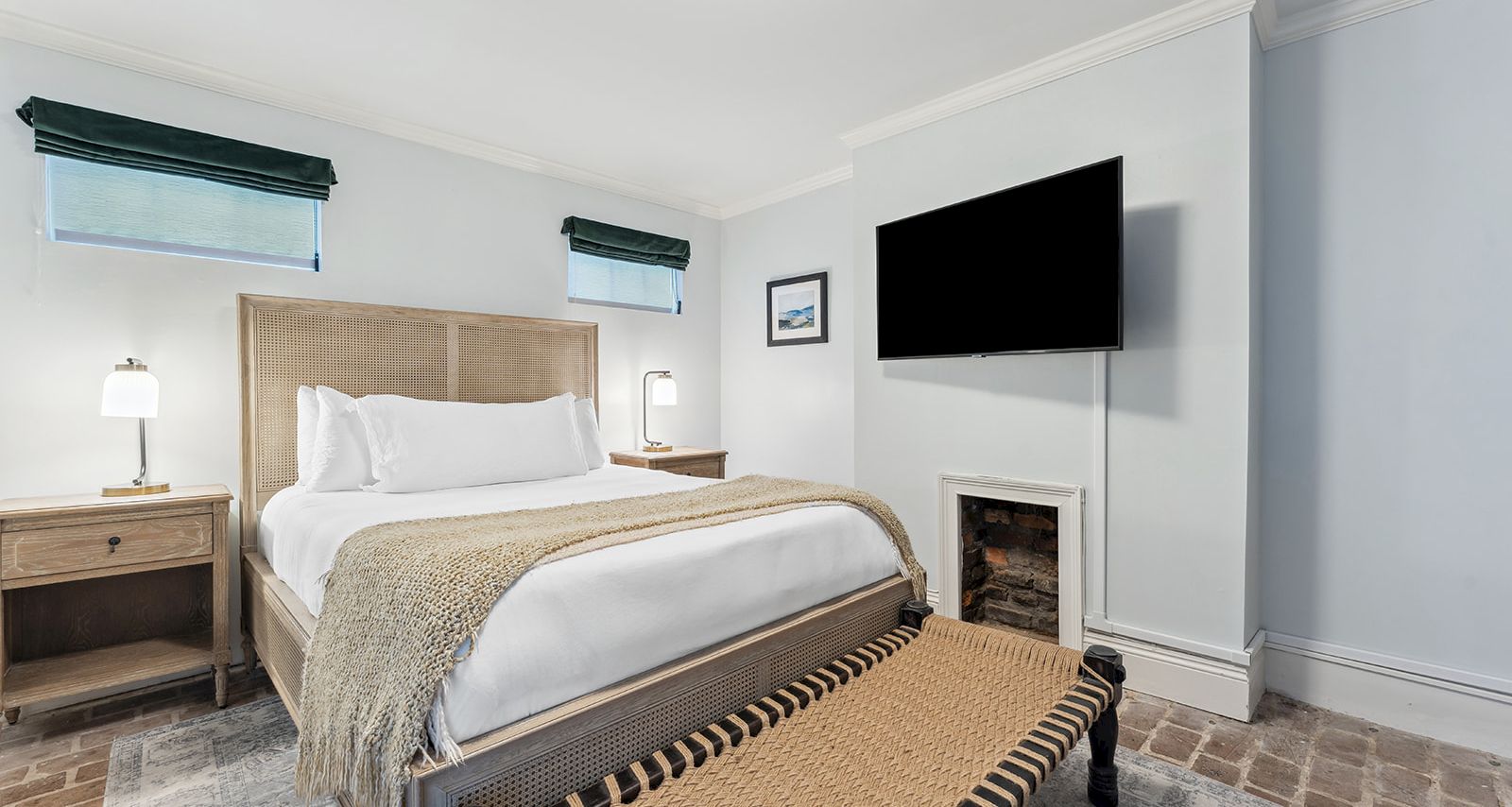 A bright hotel bedroom with a king bed, two nightstands, a bench at the foot, a wall-mounted TV, and a small fireplace-style area. End.
