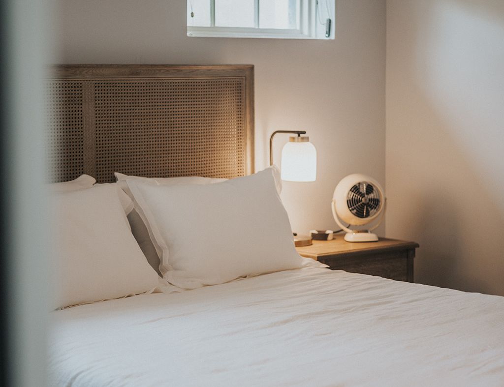 A cozy bedroom with a white bed, wicker headboard, nightstand, lamp, small window with a dark blind, and a fan on the table, all in soft neutral tones.