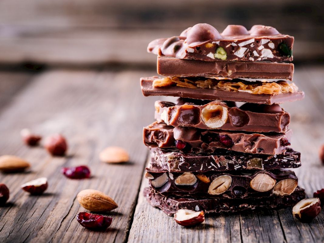 Stack of dark chocolate bars topped with nuts, dried fruit, and small candies, with crumbs on a rustic wooden table.
