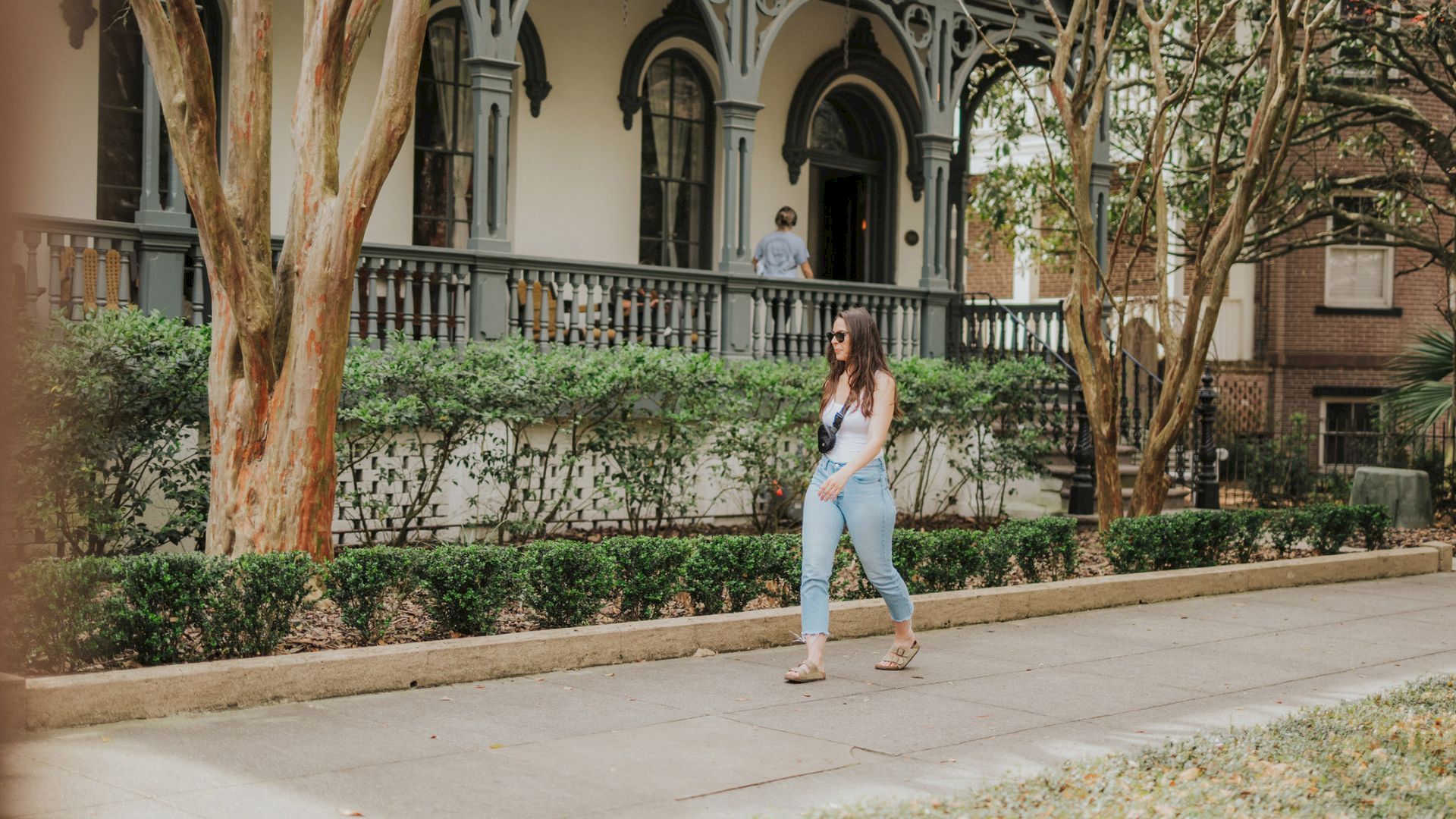 A woman in sunglasses stands on a walkway beside a historic building, with leafy hedges and trees framing the courtyard behind her.