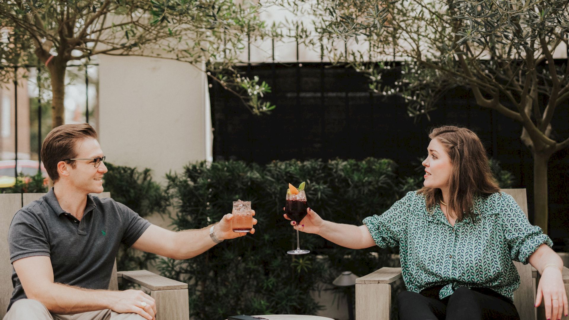 Two people on a patio cheer and clink cocktails, smiling in the garden among trees.