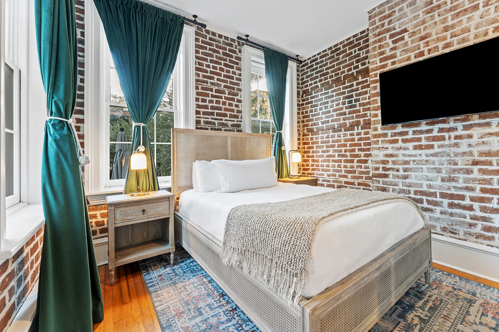A stylish brick-walled bedroom with a comfortable bed, teal curtains, two bedside tables, warm lamps, a wall-mounted TV, and a cozy rug.