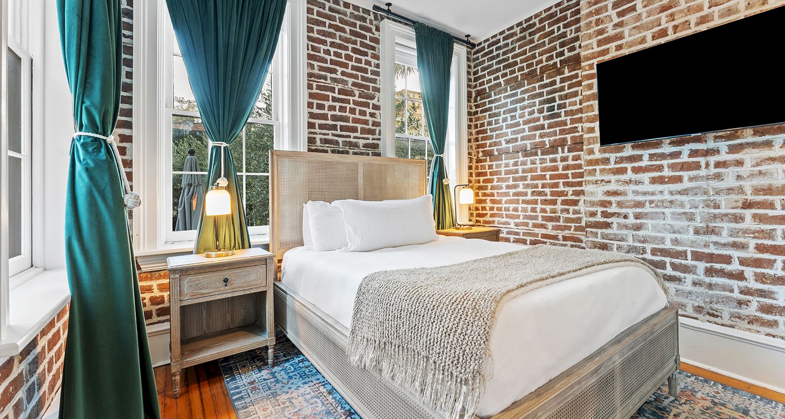 A stylish brick-walled bedroom with a comfortable bed, teal curtains, two bedside tables, warm lamps, a wall-mounted TV, and a cozy rug.